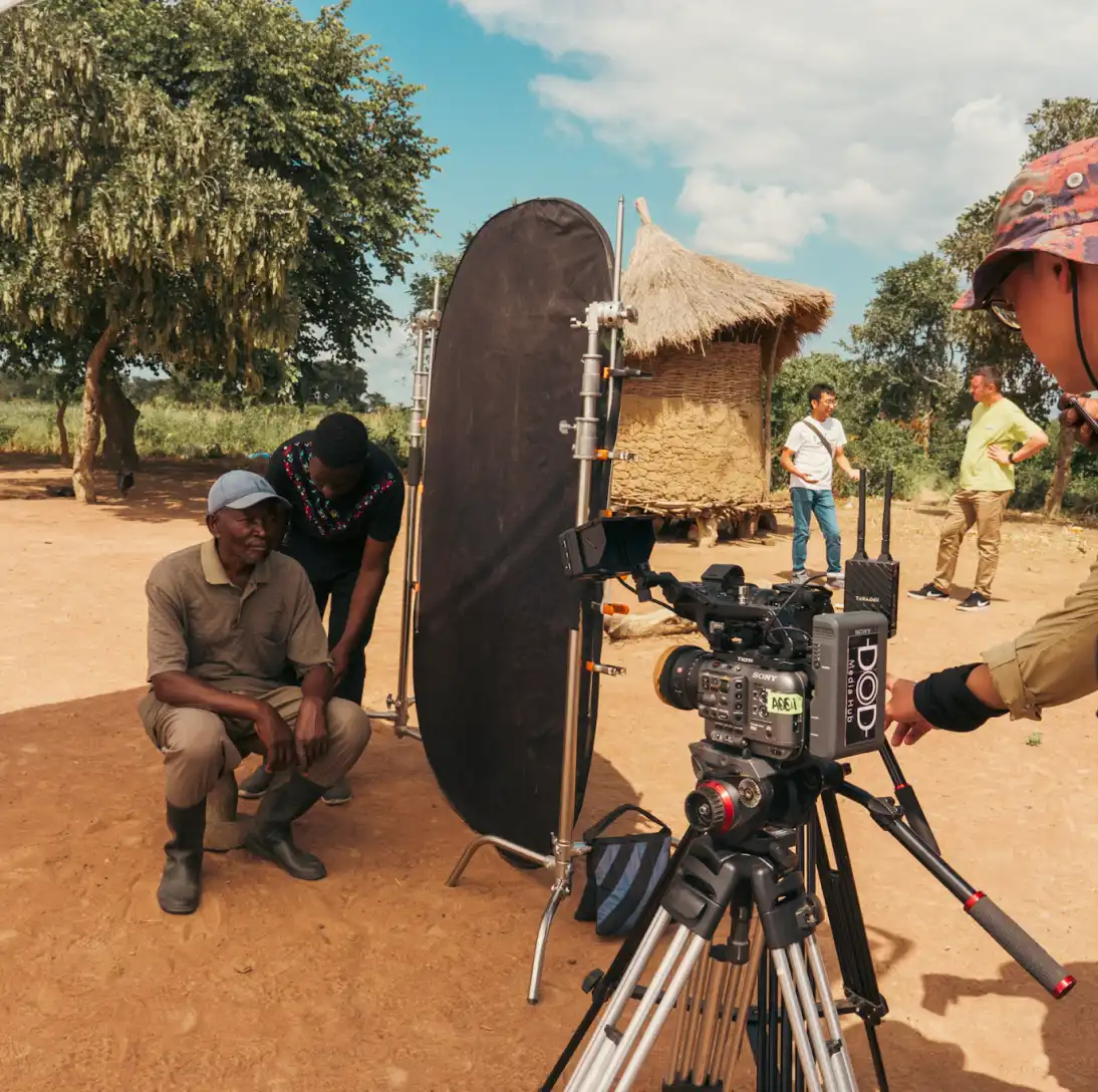 Filming crew setting up a camera outdoors with a reflector, in a rural area with traditional thatched huts.