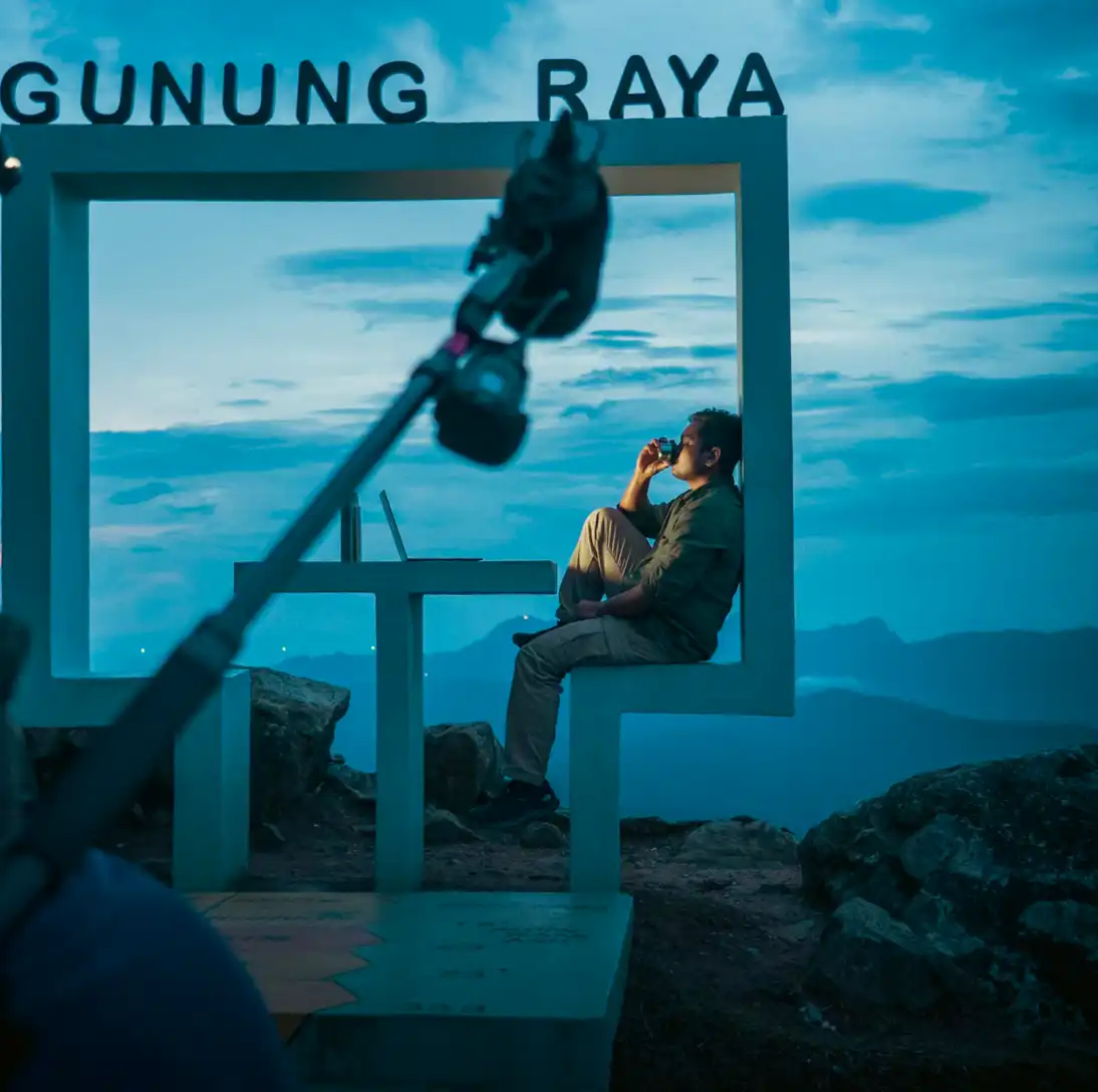 Person sitting on a bench, drinking coffee, with mountains and a blue sky in the background, during dusk.