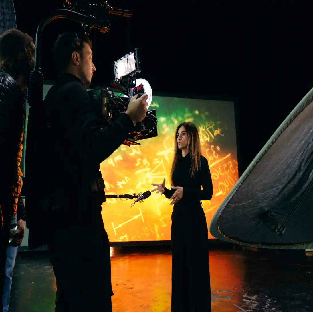Woman speaking in front of a green screen with mathematics equations, while a cameraman films her.