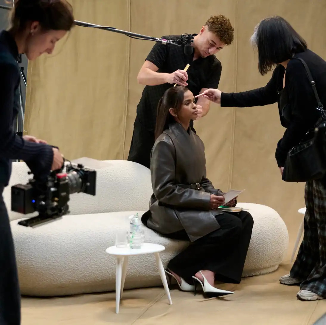 Woman sitting on a sofa during a film or photo shoot with a makeup artist and camera crew.