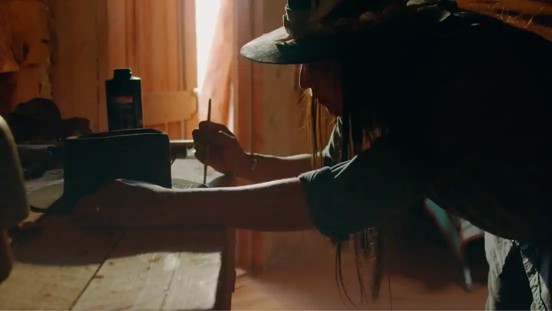 Woman handcrafting jewellery in a wooden workshop, wearing a wide-brimmed hat.