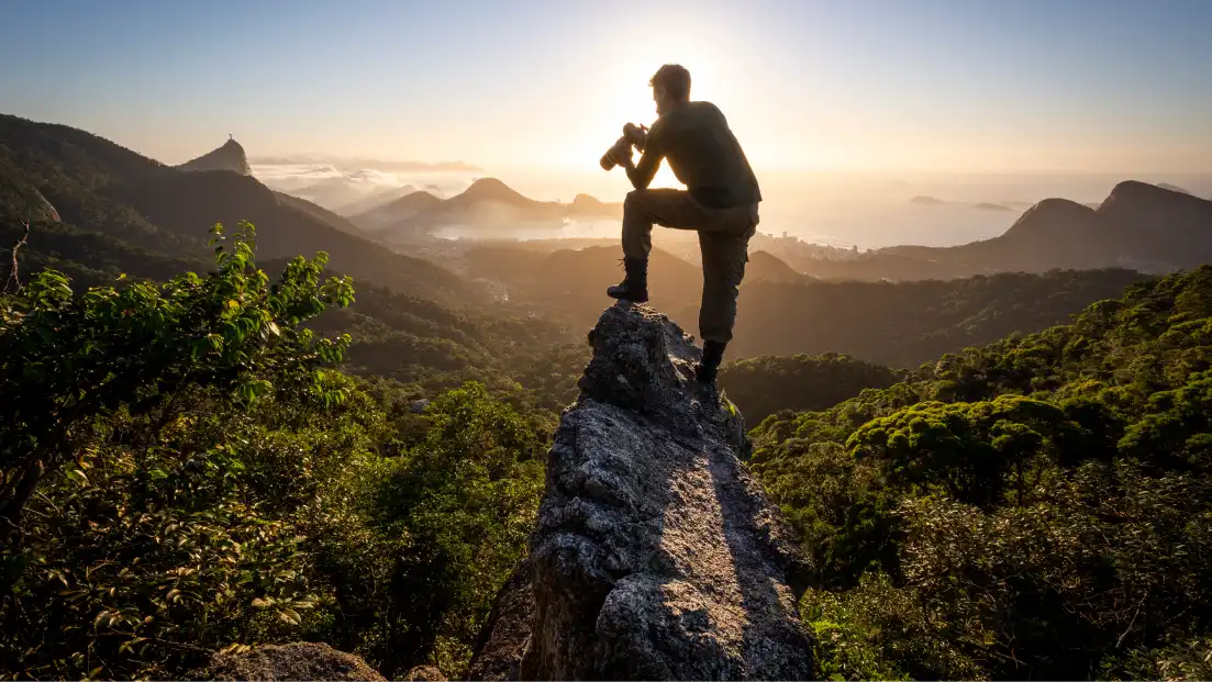 Person sitting on a rock with a camera, overlooking a mountain landscape at sunrise