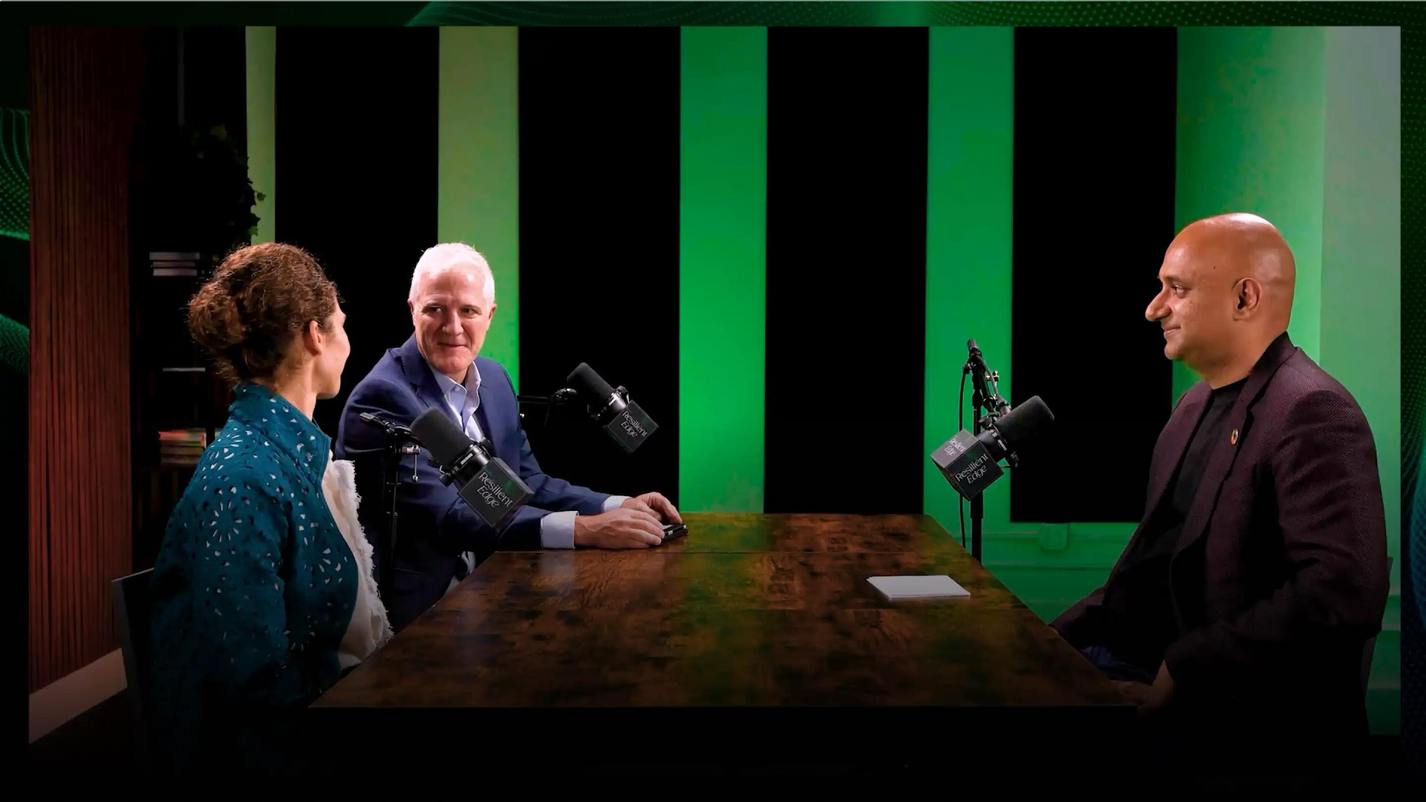 Three people seated at a table with microphones, in a recording studio with green and black acoustic panels.
