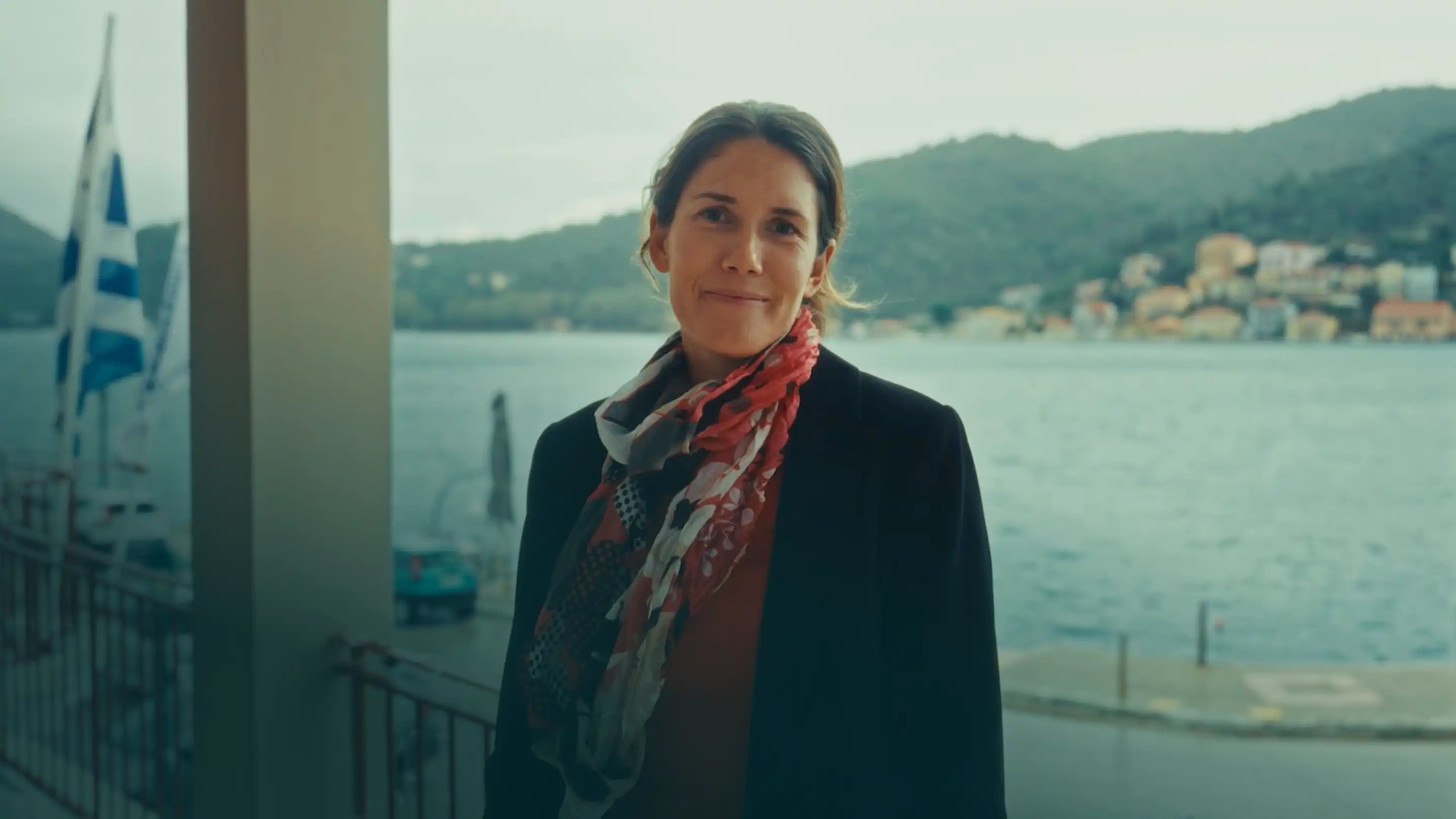 Woman standing by a waterfront with boats and hills in the background