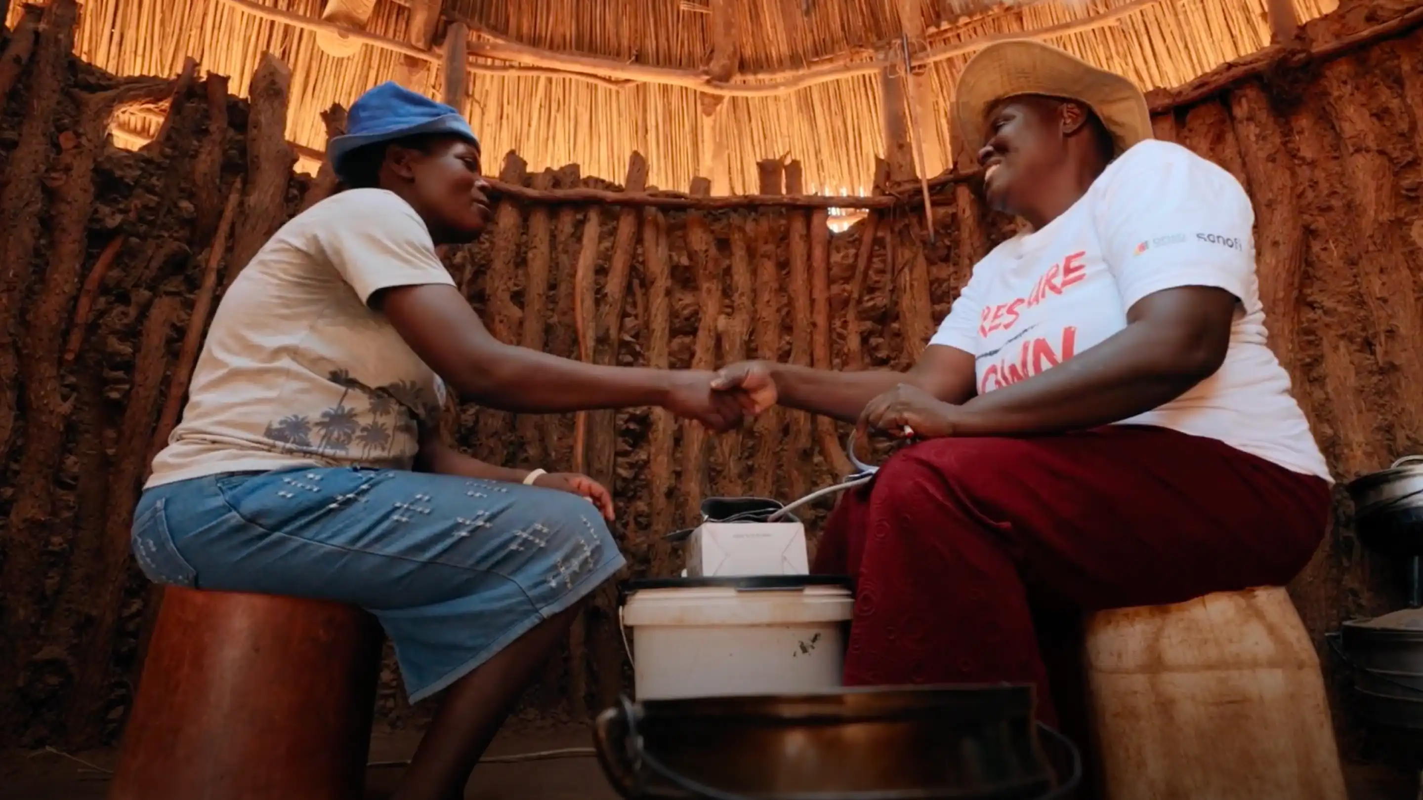 Two women shaking hands inside a thatched hut, one sitting on a wooden stool and the other on a plastic container.