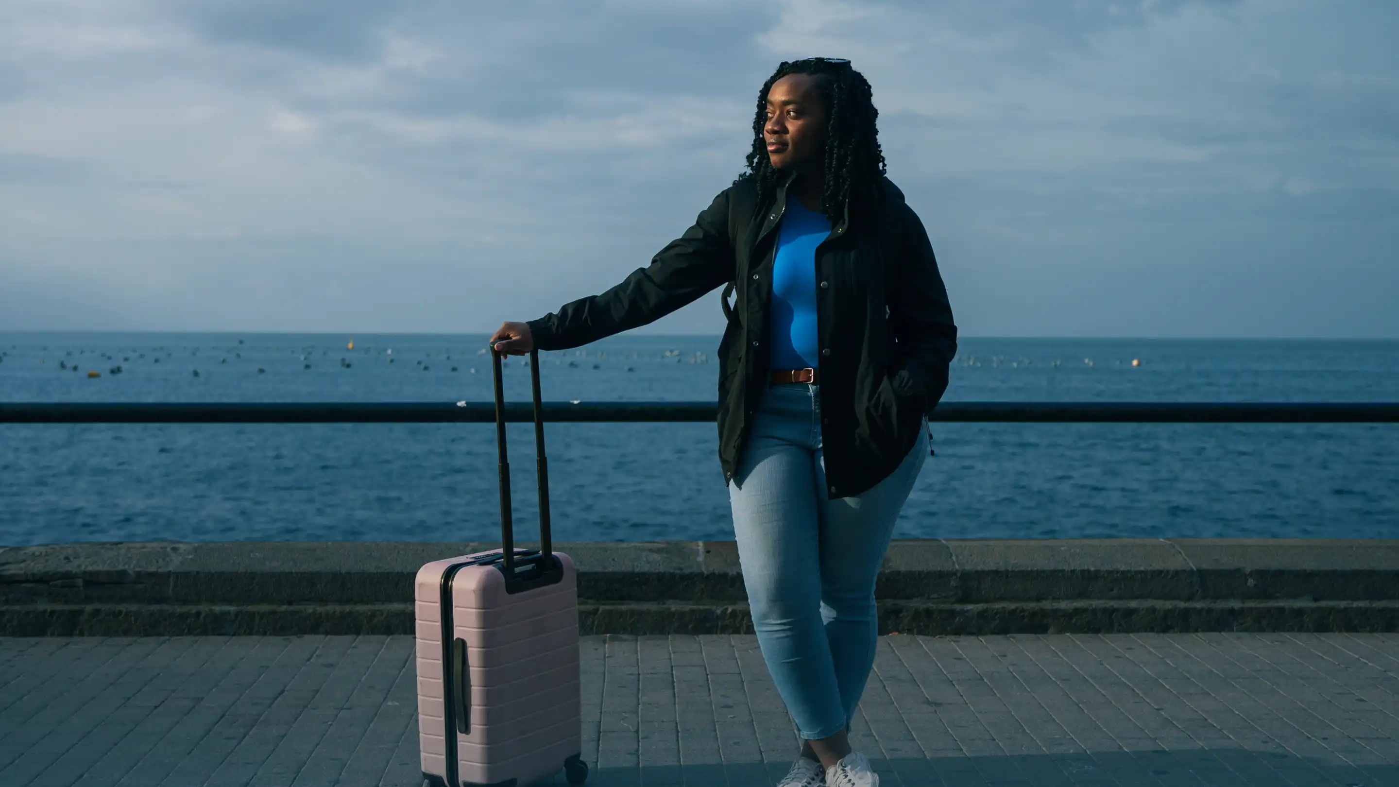 Woman with a pink suitcase standing by a waterfront railing on a cloudy day.