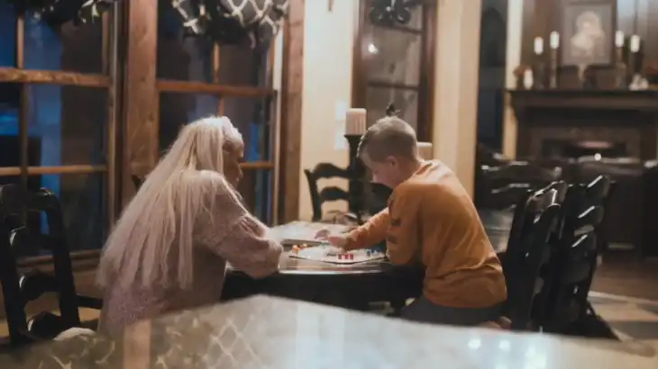 An elderly woman with long white hair and a young boy in a yellow shirt playing a board game at a dark wooden dining table.