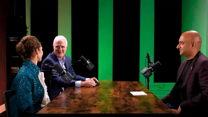 Three people seated at a table with microphones, in a recording studio with green and black acoustic panels.