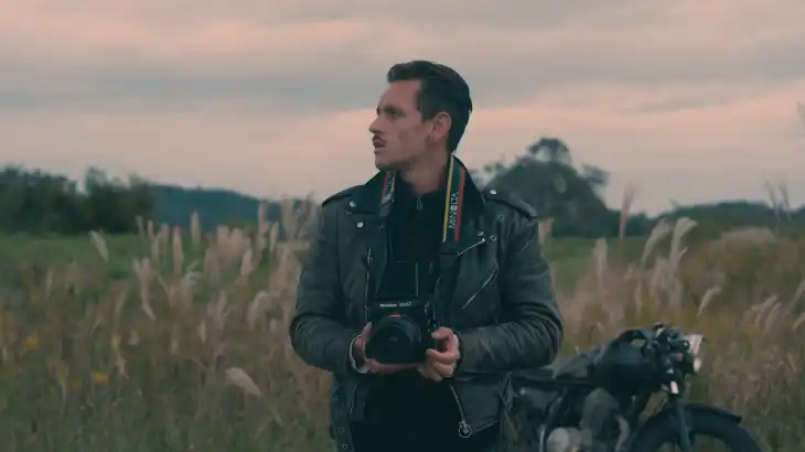 Man with a camera standing in a grassy field during sunset.