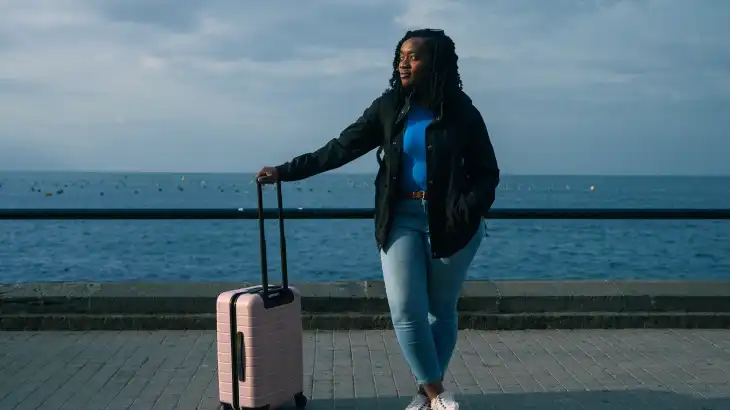 Woman with a pink suitcase standing by a waterfront railing on a cloudy day.