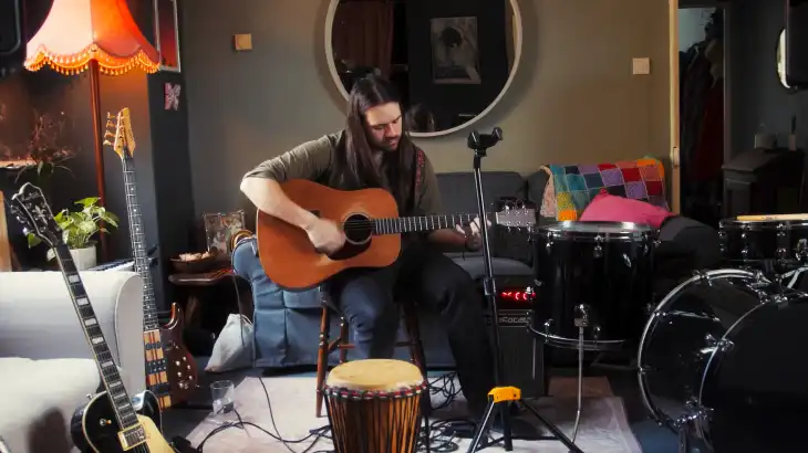 Man playing acoustic guitar in a cozy, decorated room with musical instruments.